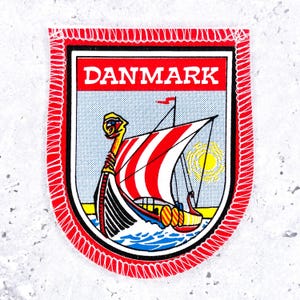 May include: A shield-shaped embroidered patch featuring a Viking ship with a red and white striped sail on a light blue background. The word DANMARK is printed in white capital letters on a red banner at the top. The patch has a red stitched border.