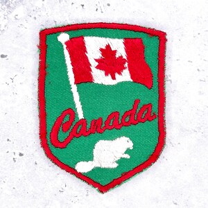 May include: A shield-shaped embroidered patch with a green background and a red border. It features a Canadian flag, the word Canada in red cursive script, and a white beaver silhouette. The design is stitched with detailed thread work.