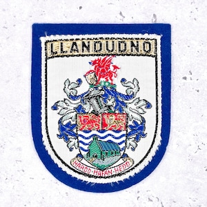 Op de afbeelding: Geborduurde patch met het woord "LLANDUDNO" bovenaan. De patch heeft een gedetailleerd wapenschild met een rode draak, een schild met een groen gebouw en de tekst "HAROD HAFAN HEDD". De patch heeft een blauwe rand.