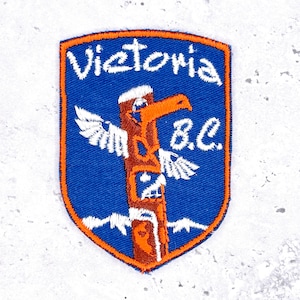 May include: Embroidered patch with a blue background and an orange border. The patch features a totem pole design with the word "Victoria" above and "B.C." below. The totem pole is brown and orange.