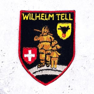 May include: A shield-shaped embroidered patch with a black background and red border. It features the yellow text "WILHELM TELL" at the top, a golden figure with a staff, a small yellow shield with a black eagle, and a red shield with a white cross.