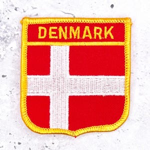 May include: A rectangular embroidered patch featuring the flag of Denmark with a red background and a white Nordic cross. The word DENMARK is stitched in yellow capital letters across the top section, bordered by a thick yellow stitched edge.