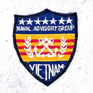 Pode incluir: Um emblema bordado em forma de escudo com seções superior e inferior azuis, estrelas brancas e o texto NAVAL ADVISORY GROUP e VIETNAM. O centro possui listras horizontais amarelas e vermelhas com um emblema branco de duas âncoras cruzadas.