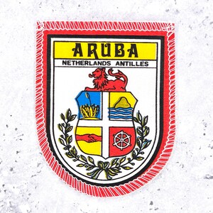May include: A shield-shaped embroidered patch featuring the text ARUBA and NETHERLANDS ANTILLES. The design includes a red lion above a quartered shield with symbols of a plant, a mountain, a handshake, and a wheel, all framed by green laurel branches.