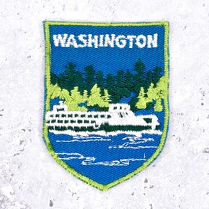 Op de afbeelding: Geborduurde patch met een blauwe achtergrond en een groene rand. De patch toont het woord "WASHINGTON" in het wit, boven een witte veerboot op blauw water en groene en lichtgroene bomen.