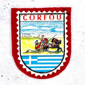 May include: A shield-shaped patch with a red felt border and a scalloped edge. The centre features the text CORFOU and VICTORY OF ACHILLES above an illustration of a chariot pulled by a black horse. The bottom section displays the blue and white Greek flag.