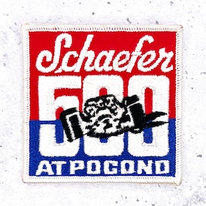 Può includere: Toppa ricamata con sfondo rosso e blu. La toppa presenta il testo "Schaefer 500" in bianco, con una grafica di auto da corsa in bianco e nero. Il testo "AT POCONO" è in basso.