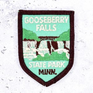 May include: A shield-shaped embroidered patch with a dark brown border. The centre features a light green background depicting Gooseberry Falls with a brown bridge, white water, and green hills. The text reads GOOSEBERRY FALLS STATE PARK MINN.