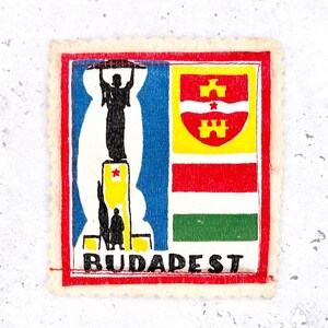 May include: A vintage postage stamp featuring a colorful design. The stamp has a red border and a cream-colored, scalloped edge. The design includes a silhouette of a statue, a coat of arms, and the word "BUDAPEST".