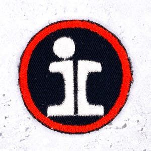 May include: A circular embroidered patch featuring a black background with a bright red border. In the center, a white stylized graphic design consists of a small circle positioned above two symmetrical, blocky shapes that resemble the letters i and c.