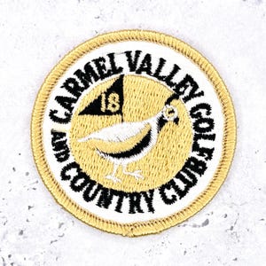 Op de afbeelding: Een ronde geborduurde patch met een geel midden, een zwarte kwartelafbeelding en een zwarte vlag met het getal 18. De witte rand bevat de zwart geborduurde tekst CARMEL VALLEY GOLF AND COUNTRY CLUB in een cirkelvormige lay-out.