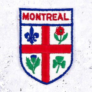 May include: A shield-shaped embroidered patch with a white background and a blue border. The word MONTREAL is stitched in red at the top. A red cross divides the shield into four quadrants containing a blue fleur-de-lis, a red rose, a green thistle, and a green shamrock.