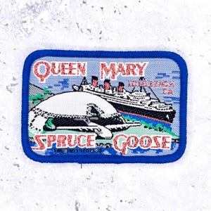 Op de afbeelding: Een rechthoekige geborduurde patch met een blauwe rand met het schip Queen Mary en het vliegtuig Spruce Goose. De tekst luidt QUEEN MARY LONG BEACH CA en SPRUCE GOOSE met de datum 1984. Het ontwerp gebruikt rood, wit en blauw.