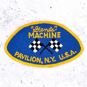Può includere: Una toppa ricamata di forma ovale con sfondo blu e bordo cucito giallo. Presenta il testo "Gene's" MACHINE e PAVILION, N.Y. U.S.A. in filo giallo, centrato con due bandiere da corsa a scacchi bianche e nere incrociate.