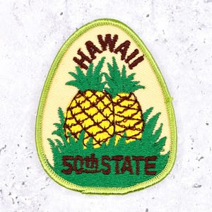 Könnte beinhalten: Ein Aufnäher mit zwei gelben Ananas mit grünen Blättern und Gras, mit dem Wort "HAWAII" darüber und "50th STATE" darunter, alles in braunem Text. Der Aufnäher hat einen hellgelben Hintergrund und einen grünen Rand.