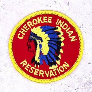 May include: A round, embroidered patch with a red background and yellow border. The patch features a profile of a Native American in a headdress with yellow, blue, and black details. The text "CHEROKEE INDIAN RESERVATION" is embroidered around the edge.