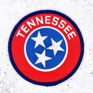 Op de afbeelding: Een cirkelvormige geborduurde patch met het ontwerp van de vlag van de staat Tennessee. De patch heeft een blauwe centrale cirkel met drie witte sterren, omringd door een rode rand met het woord TENNESSEE in witte hoofdletters en een donkerblauwe rand.