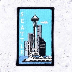 May include: Embroidered patch featuring the Seattle skyline, including the Space Needle and other buildings, set against a light blue background. The word "SEATTLE" is vertically embroidered on the left side. The patch has a black border.