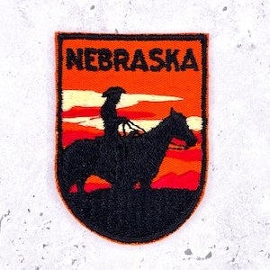 May include: Embroidered patch with the word "NEBRASKA" above a silhouette of a rider on horseback against a sunset. The patch has an orange background with red and yellow accents, and a black border.