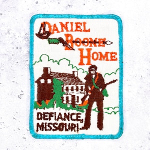 May include: A rectangular embroidered patch with a bright turquoise border on a white background. It features the orange text DANIEL BOONE HOME, a brown house, green foliage, and a brown figure holding a rifle, with DEFIANCE, MISSOURI below.
