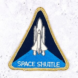 Può includere: Una toppa ricamata triangolare con la grafica di uno space shuttle bianco su sfondo blu scuro. La sezione inferiore è azzurra con la scritta SPACE SHUTTLE in bianco. L'intera toppa è rifinita con un bordo in filo dorato metallizzato.