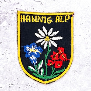 Könnte beinhalten: Ein schwarzer, gestickter Aufnäher mit dem Text "HANNNIG ALP" in Gelb. Der Aufnäher zeigt eine weiße Gänseblümchen, eine blaue Blume und rote Blumen mit grünen Stielen und Blättern, alle gelb umrandet.