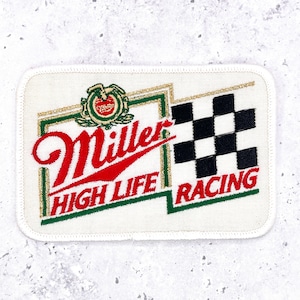 Pode incluir: Um emblema bordado retangular com fundo branco e borda costurada. Apresenta o logótipo vermelho Miller High Life, um emblema verde e dourado e um padrão de bandeira axadrezada a preto e branco com a palavra RACING em letras vermelhas.
