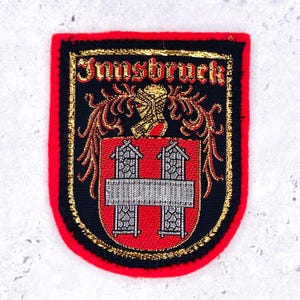 Op de afbeelding: Een rechthoekige geborduurde patch met een felrode rand. Het midden toont een zwart schild met het woord Innsbruck in rode tekst, een gouden helm als helmteken en een rood wapenschild met twee zilveren torens verbonden door een brug.
