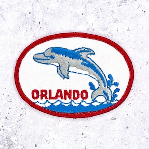 May include: Oval patch featuring a leaping dolphin in blue and grey, above blue waves. The word "ORLANDO" is in red, below the dolphin. The patch has a red border and a white background.