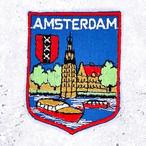 Pode incluir: Patch bordado com fundo azul e borda vermelha. O patch apresenta a palavra "AMSTERDAM" no topo, um escudo com três cruzes pretas e uma representação da arquitetura e barcos de Amsterdã. O patch é uma lembrança.