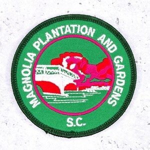 May include: A round, green patch with the text "MAGNOLIA PLANTATION AND GARDENS" in white, encircling a pink ring. The center features a white bridge and red and pink floral imagery. The text "S.C." is at the bottom.