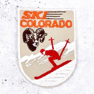 Op de afbeelding: Een rechthoekige geborduurde patch met een beige achtergrond en de oranje tekst "SKI COLORADO" bovenaan. Onder de tekst staat een bruine kop van een dikhoornschaap en een figuur in een rode outfit die een witte helling afskiet met rode stokken.