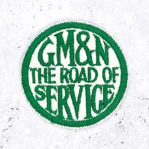 May include: A circular embroidered patch featuring a white background with a thick green border. The text GM&N THE ROAD OF SERVICE is stitched in green thread in a bold, stylised font across the centre of the circular patch design.