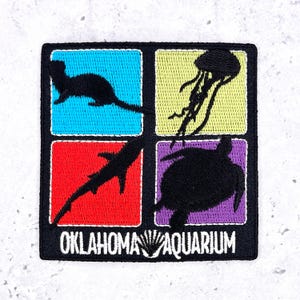 May include: A square embroidered patch featuring four colorful quadrants with black silhouettes of an otter, a jellyfish, a shark, and a sea turtle. The text OKLAHOMA AQUARIUM is embroidered in white at the bottom against a black border.