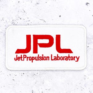 May include: A rectangular white embroidered patch with rounded corners featuring the red text JPL in a bold, stylized font above the red text Jet Propulsion Laboratory in a smaller, sans-serif font, all stitched onto a white fabric base.