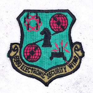 May include: Embroidered patch with a teal background and a black border. The patch features a black chess piece, two red and black globes, and a red geometric shape. The text "6950 ELECTRONIC SECURITY WING" is in gold.