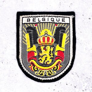 Op de afbeelding: Geborduurde patch met het woord "BELGIQUE" bovenaan. De patch toont een schildontwerp met een gele leeuw, een kroon en rode en gele details. De rand is zwart.
