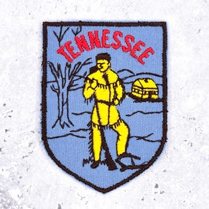 Puede incluir: Un parche bordado en forma de escudo con fondo azul claro y borde marrón oscuro. Presenta una figura amarilla con ropa de flecos, un árbol desnudo y una pequeña cabaña. La palabra TENNESSEE está bordada en letras rojas en la parte superior.