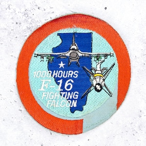 May include: A circular embroidered patch featuring a blue silhouette of the state of Illinois on a light blue background, surrounded by an orange border. It depicts an F-16 fighter jet, a missile, and the text 1000 HOURS F-16 FIGHTING FALCON.