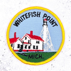 Pode incluir: Patch bordado com um farol e edifícios com telhados vermelhos e o texto "WHITEFISH POINT MICH." O patch tem um fundo azul e uma borda amarela. O farol é branco com o topo vermelho.