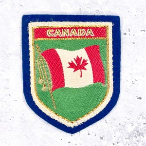 Może przedstawiać: Haftowana naszywka z flagą Kanady i napisem „CANADA” w złotym kolorze. Naszywka ma niebieską obwódkę i kształt tarczy. Flaga przedstawia czerwony liść klonu na białym tle z czerwonymi pasami.