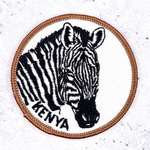 Könnte beinhalten: Runder, gestickter Aufnäher mit einem Zebrastreifenkopf in Schwarz und Weiß auf weißem Hintergrund. Der Aufnäher hat einen braunen Rand und den Schriftzug "KENYA".