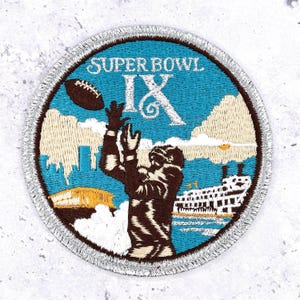 May include: Circular patch with a silver border featuring the text "SUPER BOWL IX". The patch depicts a football player catching a football, with a cityscape and a steamboat in the background. The patch is primarily blue, brown, and white.