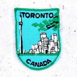 May include: Embroidered patch featuring a teal background with the words "TORONTO" and "CANADA". The patch depicts the CN Tower, a cityscape, and a green tree branch. The patch has a green border.