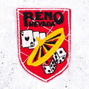 Peut inclure: Un écusson brodé en forme de bouclier avec un fond rouge et une bordure blanche. Il présente le texte noir RENO NEVADA, trois cartes à jouer blanches avec des enseignes noires, une roulette jaune et deux dés blancs avec des points noirs.