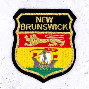May include: Embroidered patch featuring the New Brunswick provincial shield. The shield has a black border with gold stitching. The top section is black with "NEW BRUNSWICK" in gold. Below is a red section with a gold lion, and a yellow section with a ship.