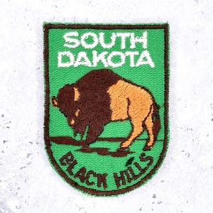 Puede incluir: Un parche bordado rectangular con fondo verde y borde marrón oscuro. Presenta un bisonte marrón y beige de pie sobre un suelo marrón oscuro. El texto SOUTH DAKOTA está bordado en blanco en la parte superior y BLACK HILLS en marrón en la parte inferior.