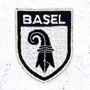 May include: A shield-shaped embroidered patch featuring a black background with a white border. The word BASEL is stitched in white capital letters at the top. Below the text, a black Basel crozier symbol is centered on a white shield field.