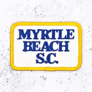 May include: Embroidered patch featuring the text "MYRTLE BEACH S.C." in blue on a white background. The patch has a yellow border and a rectangular shape. Likely a souvenir or travel memento.