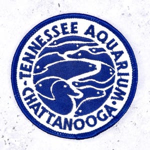 May include: A circular embroidered patch featuring a white border with the blue text TENNESSEE AQUARIUM CHATTANOOGA. The center displays a blue and white graphic of multiple fish swimming in a circular pattern, with a duck silhouette at the bottom.